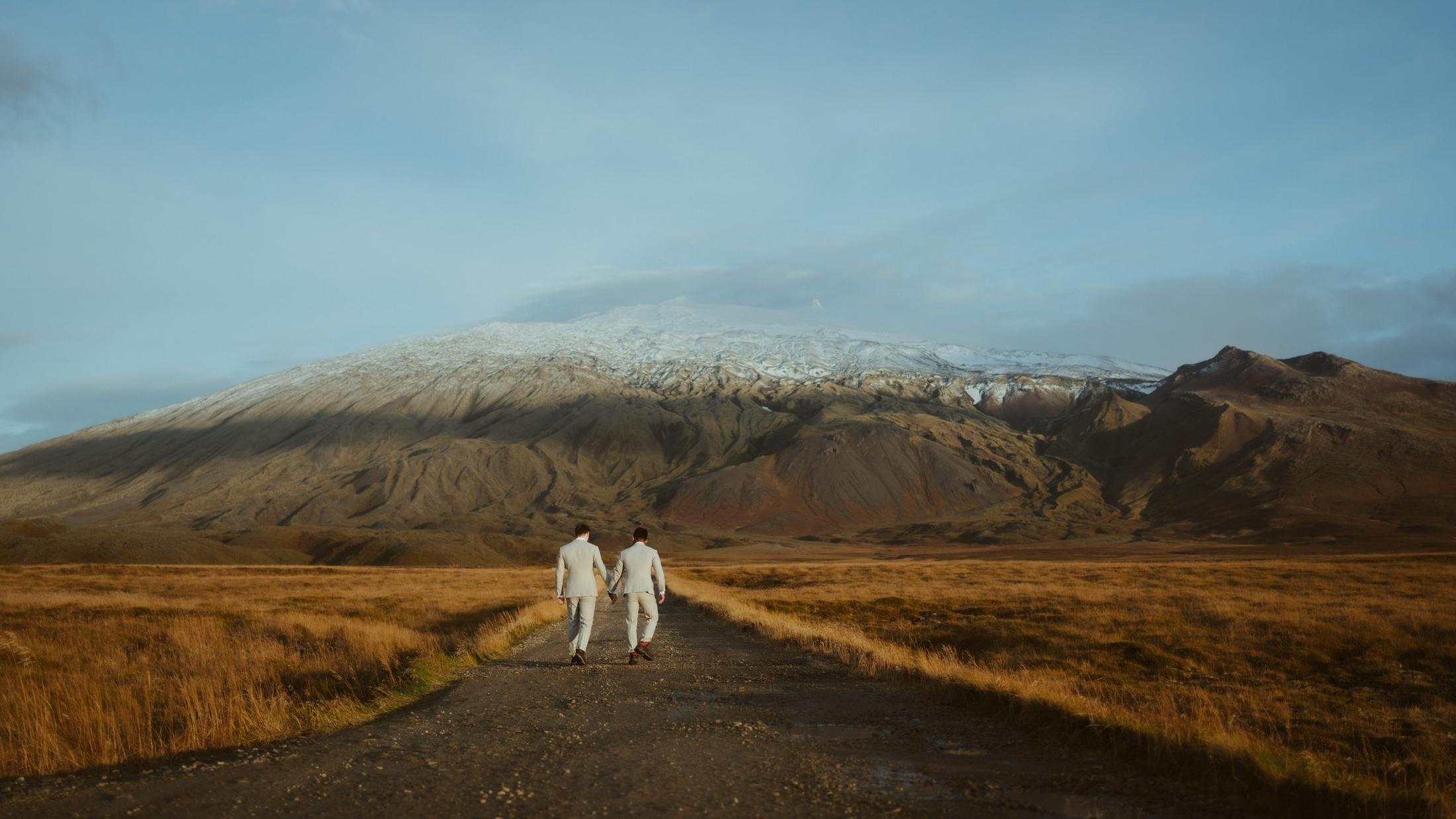 Two people walking along a road towards a snow-dusted mountain.