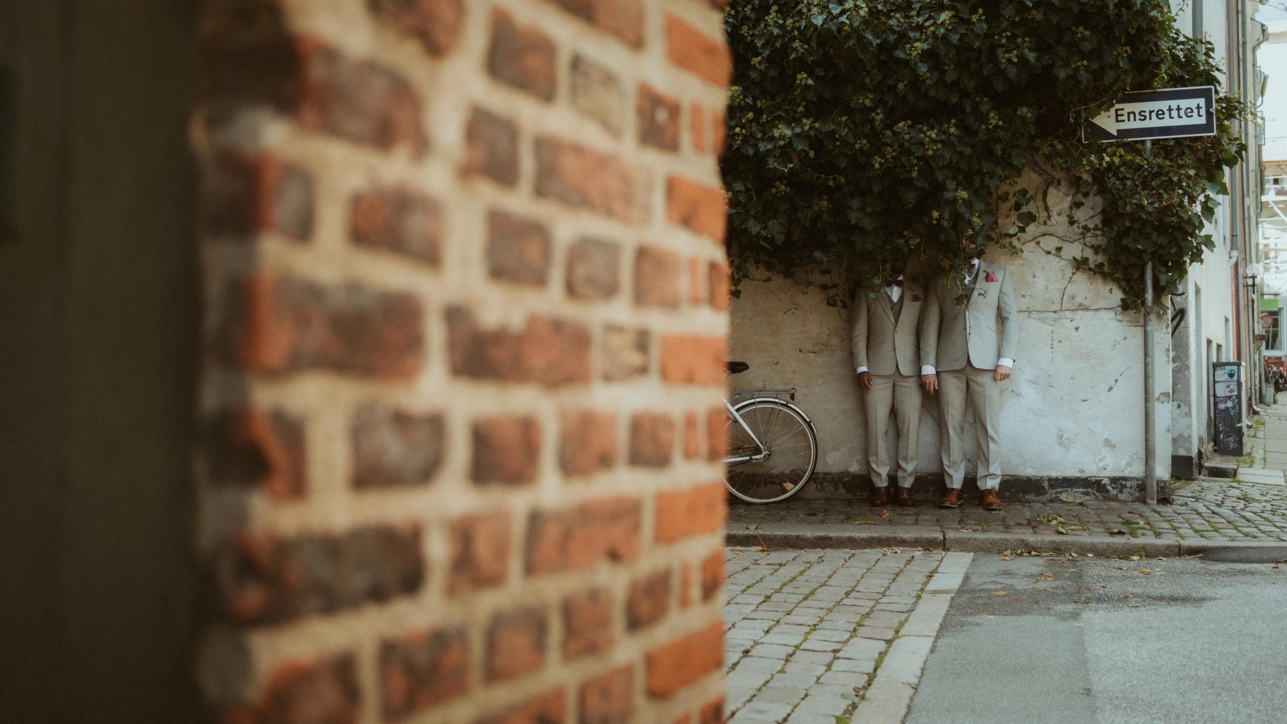 Three people in light suits standing beneath ivy beside a bicycle in Copenhagen.