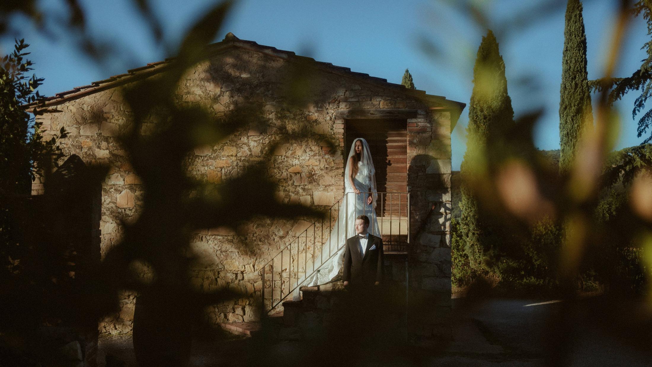 The couple stands outside a Tuscan stone house, seen through dark branches.