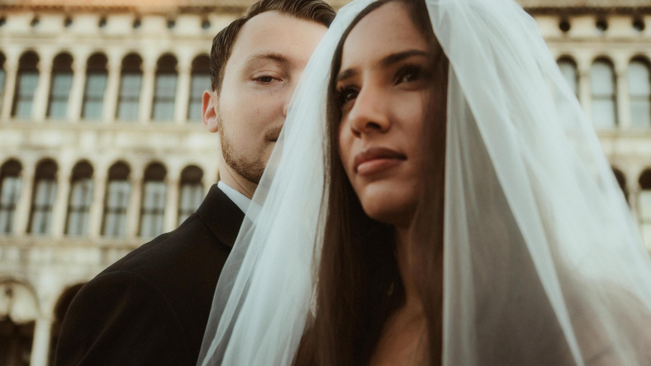 Close portrait of the bride under her veil with her partner partly visible behind her.