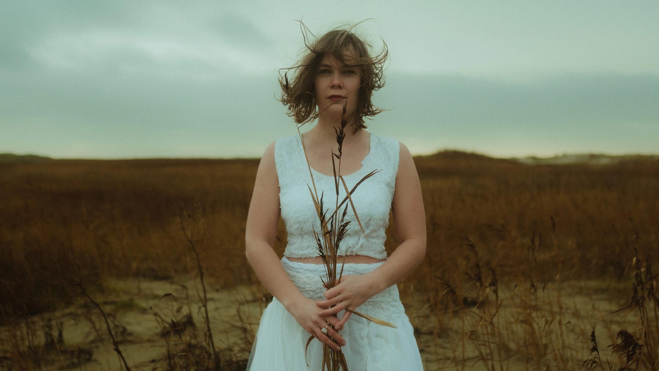 Anni stands alone holding dry reeds in front of her white dress.