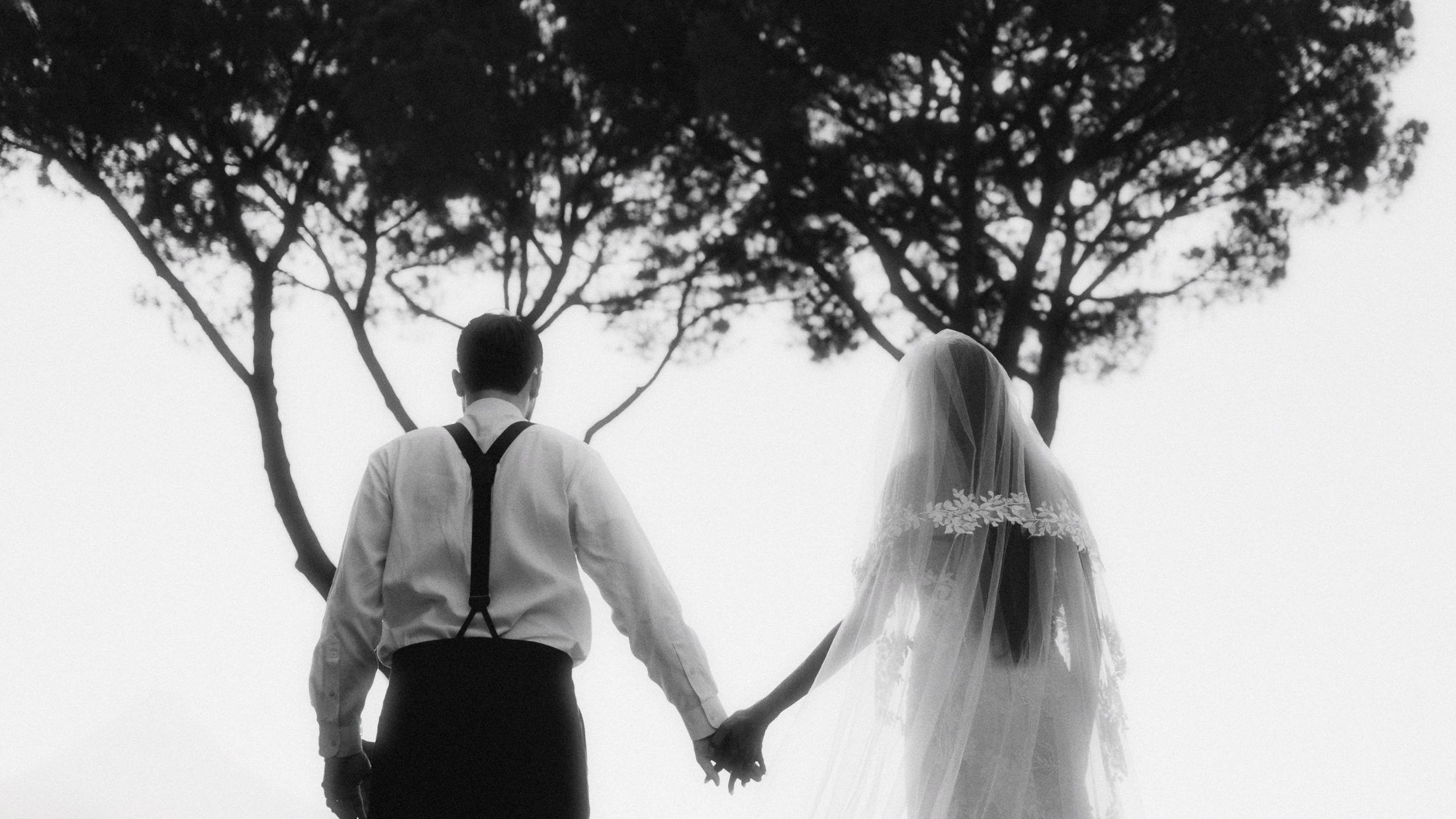 Black-and-white rear view of the couple walking hand in hand beneath tall trees.