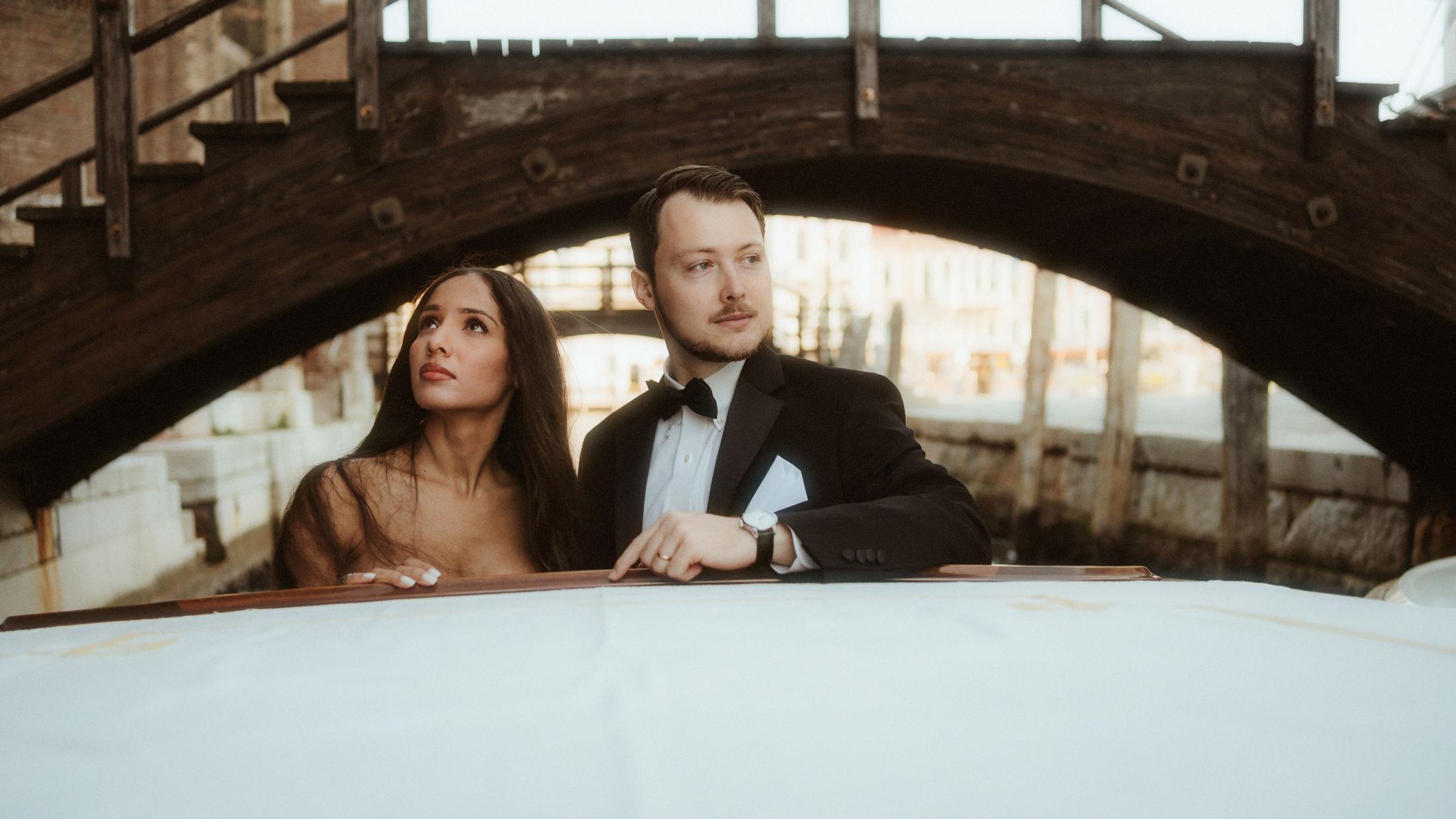 The couple looks upward together from a boat passing beneath a low bridge.