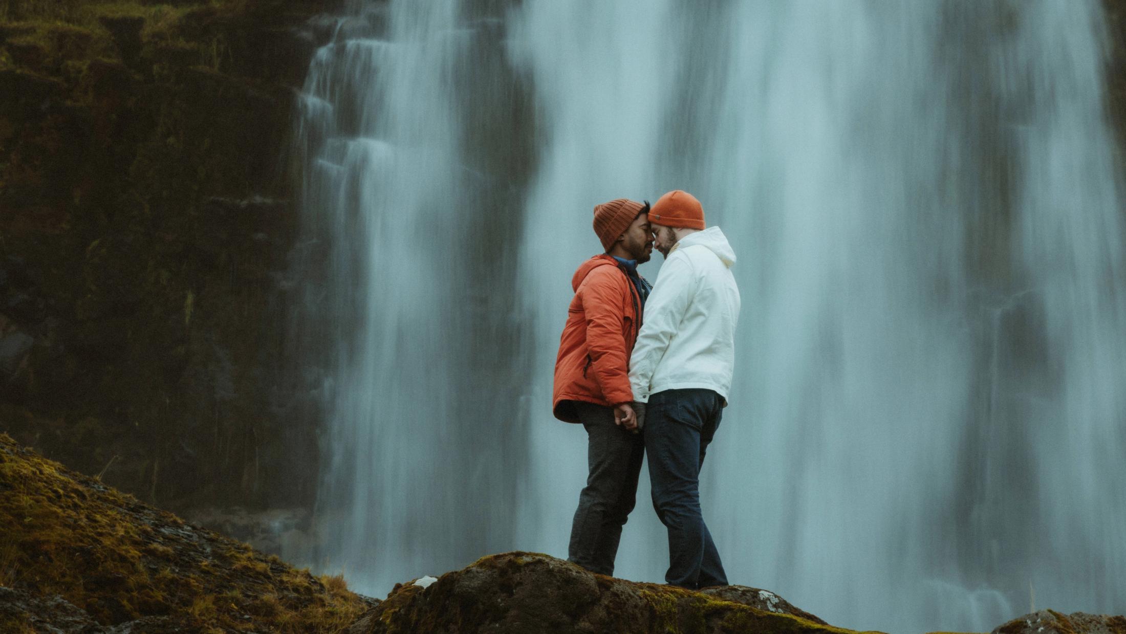 Das Paar küsst sich auf dunklen Felsen, während der Wasserfall den Hintergrund füllt.