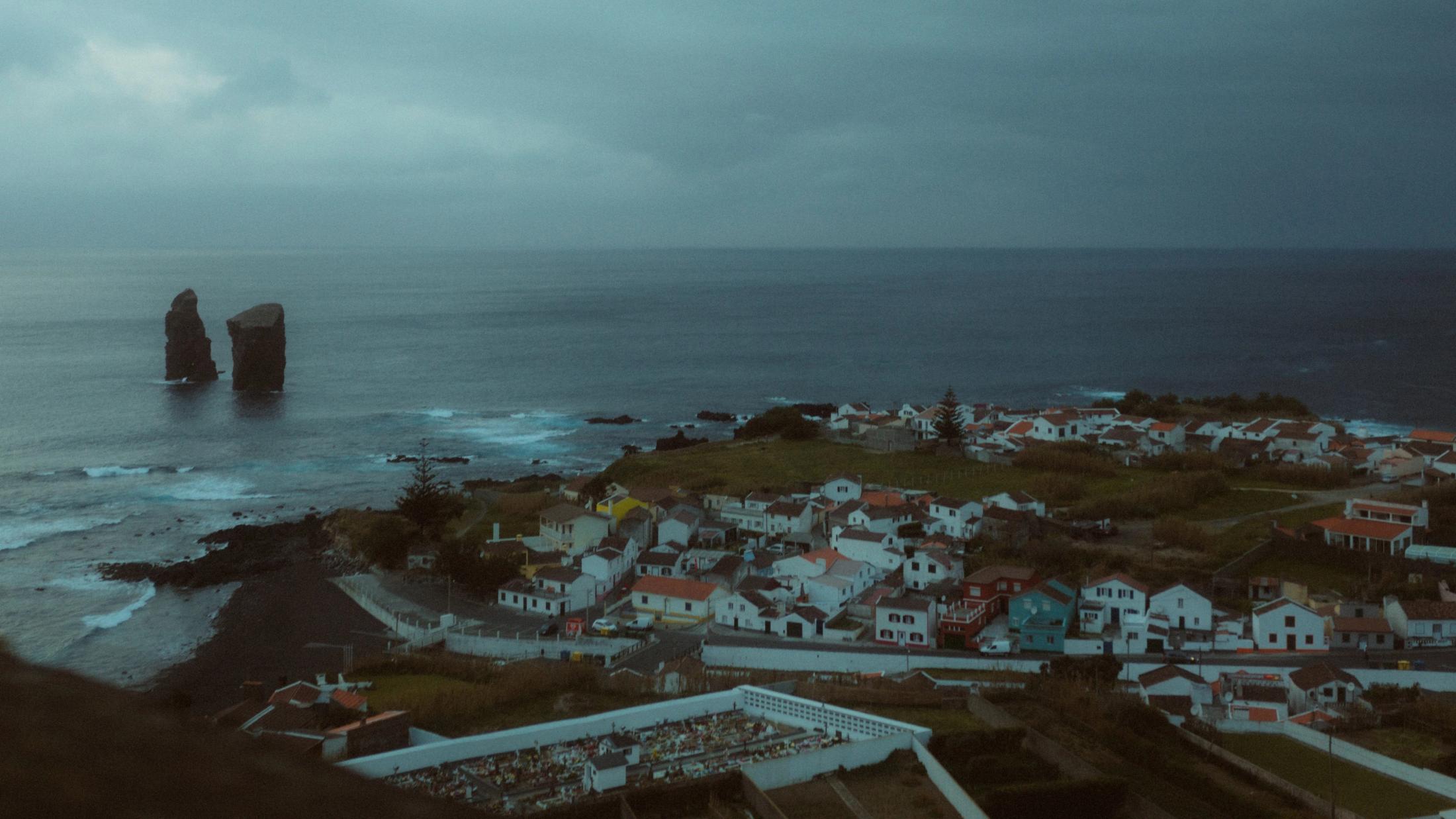 Coastal village and volcanic shoreline on São Miguel beneath a blue evening sky.
