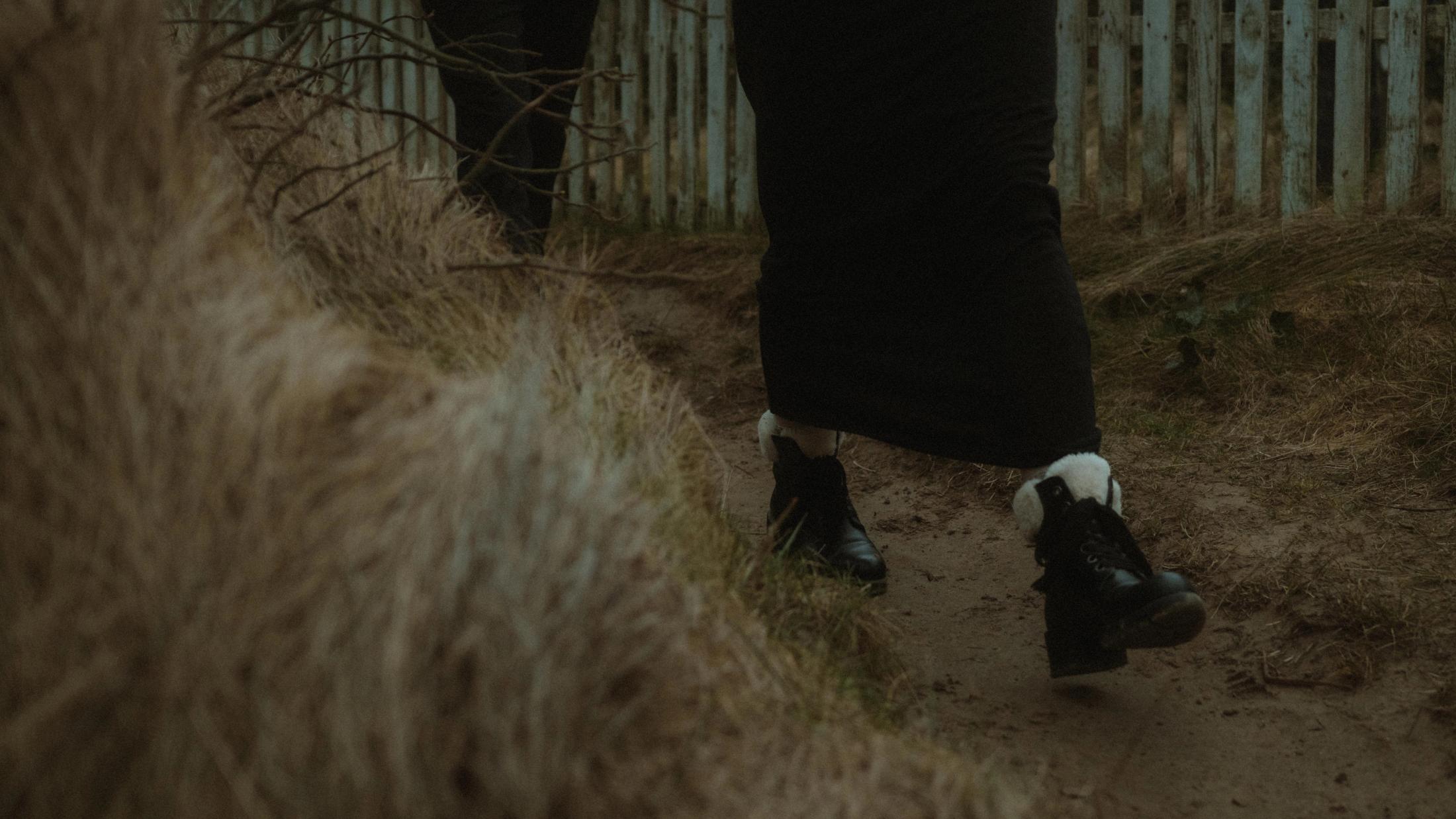 Schwarze Schuhe gehen durch trockenes Gras neben einem alten Holzzaun.