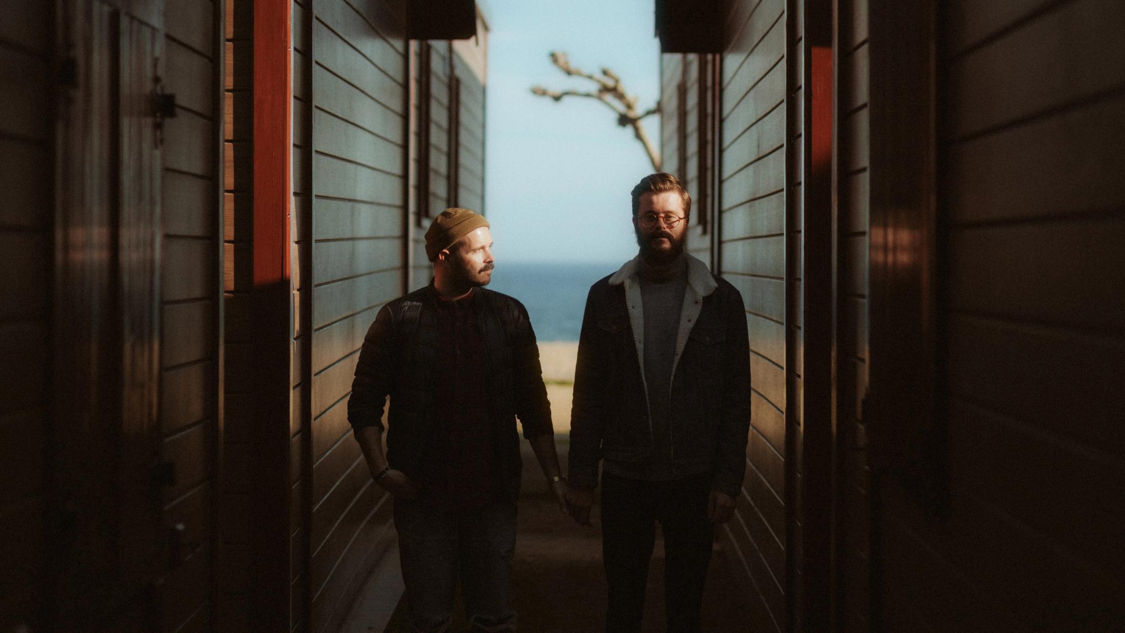Alberto and Yago walk side by side through a narrow passage between wooden houses.