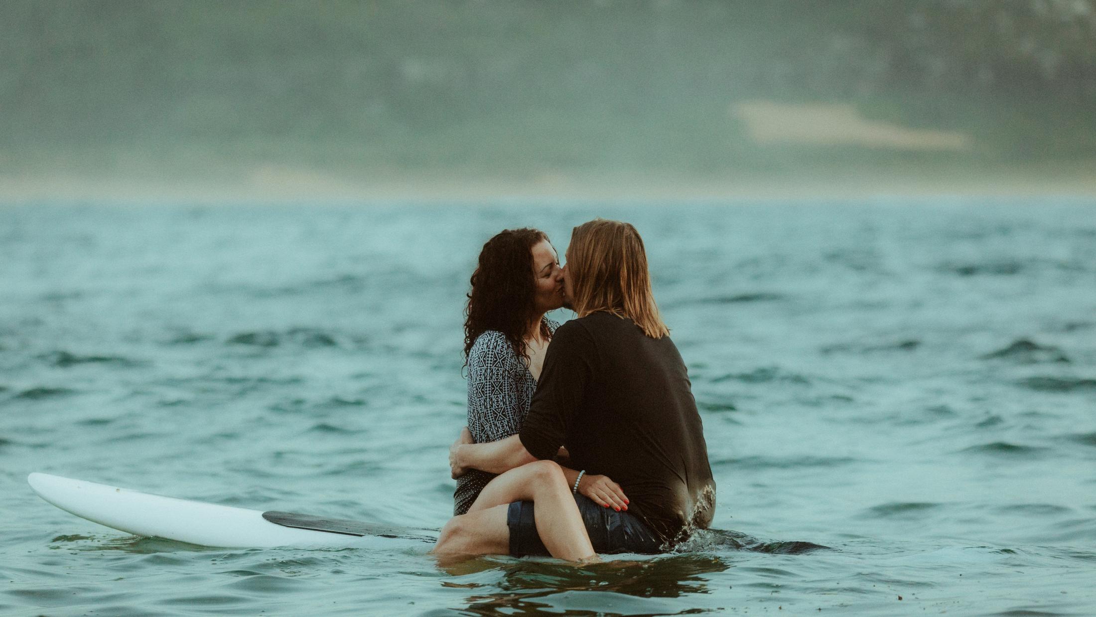 Petra and Roman holding each other on a surfboard with the coast behind them