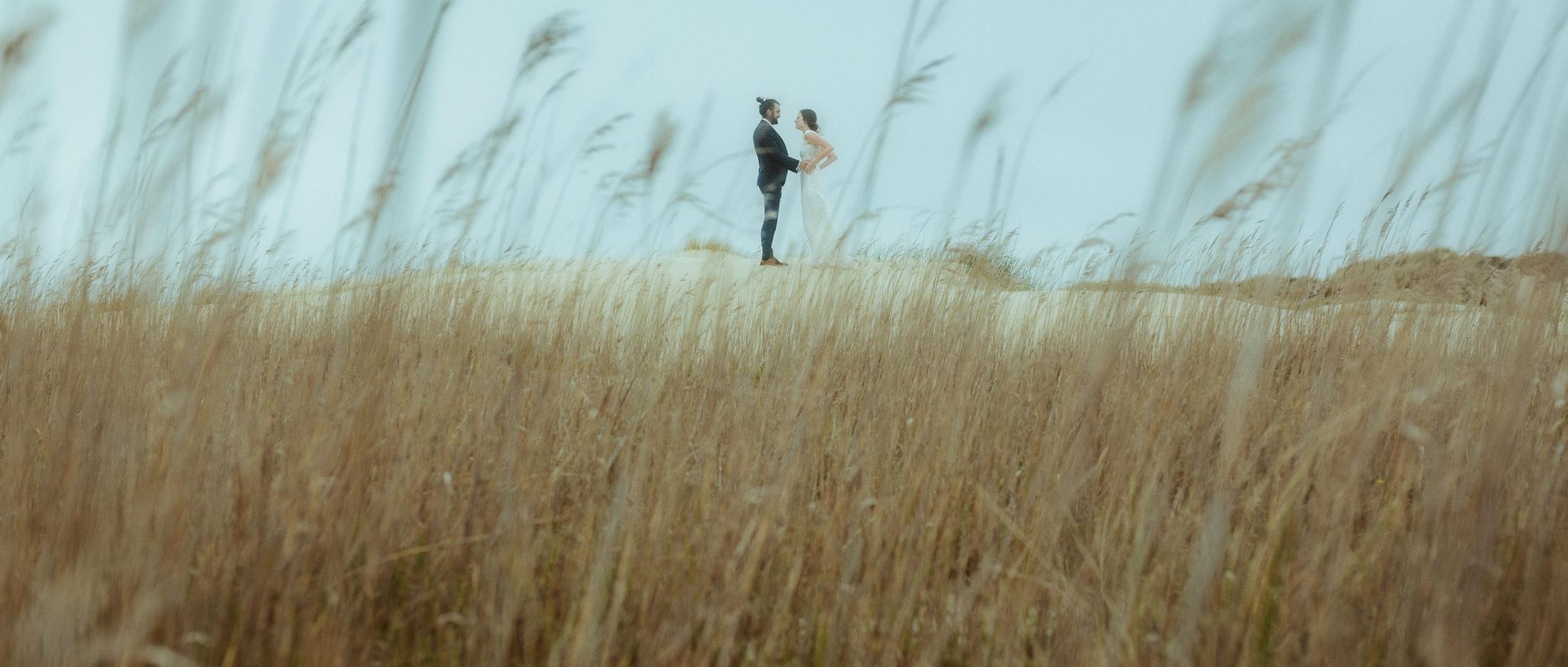Nina und Torben stehen in der Ferne hinter hohem, vom Wind bewegtem Dünengras.