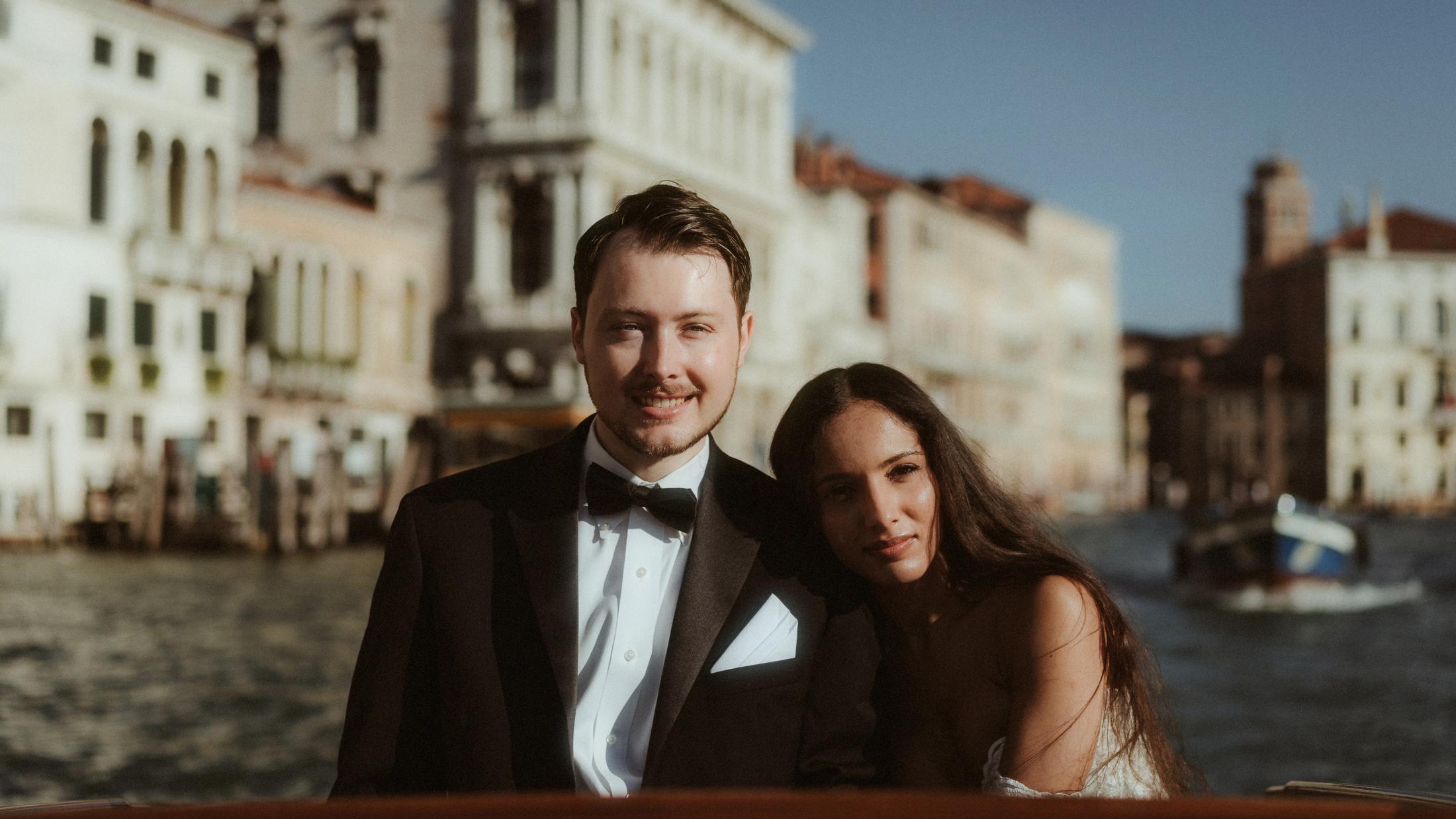 The couple sits close together on a boat with Venetian buildings behind them.