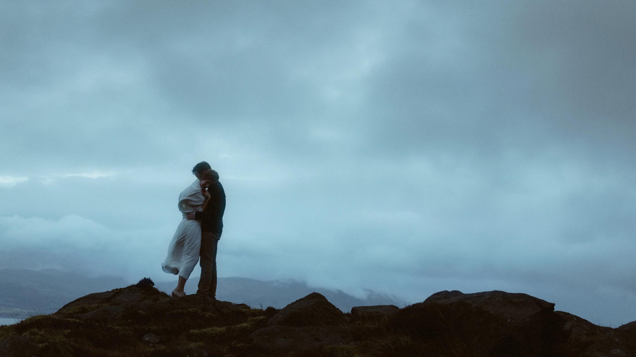 Jule und Rory umarmen sich unter offenem grauem Himmel an einem Berghang.