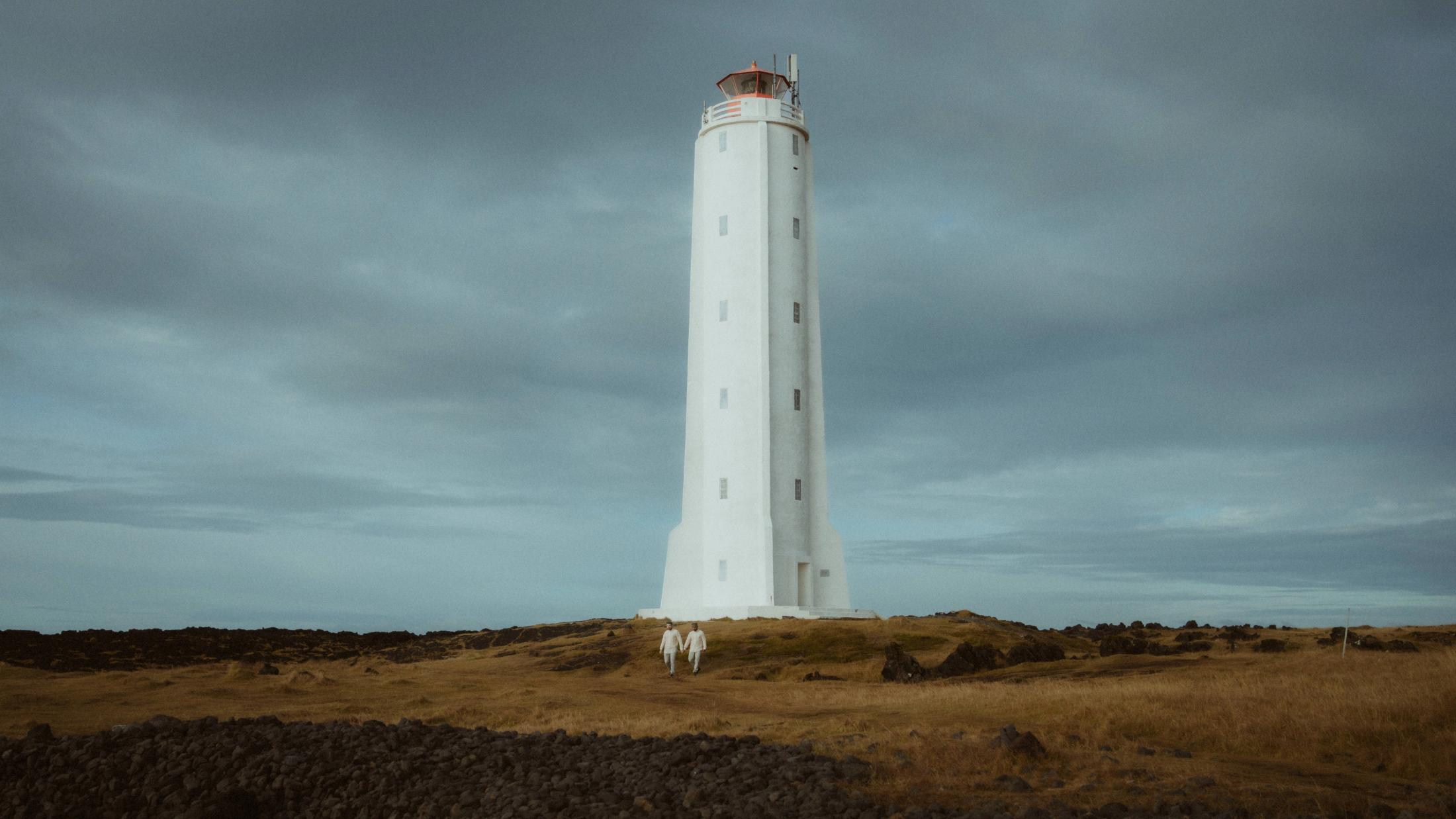 Ein weißer Leuchtturm steht hinter dem Paar in einem weiten Feld unter grauen Wolken.