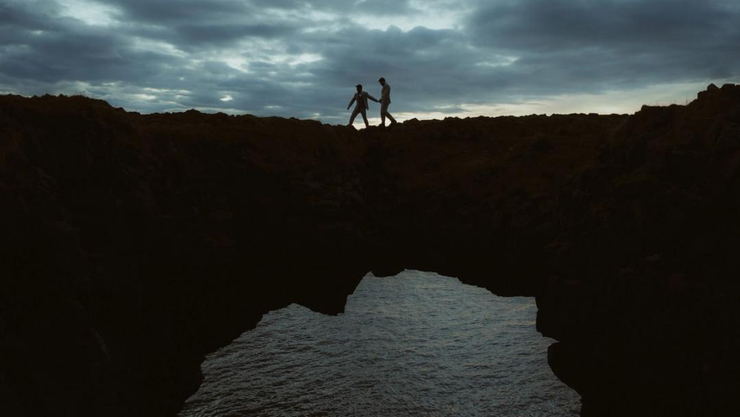 Two people holding hands on a rock arch above the sea in Iceland.