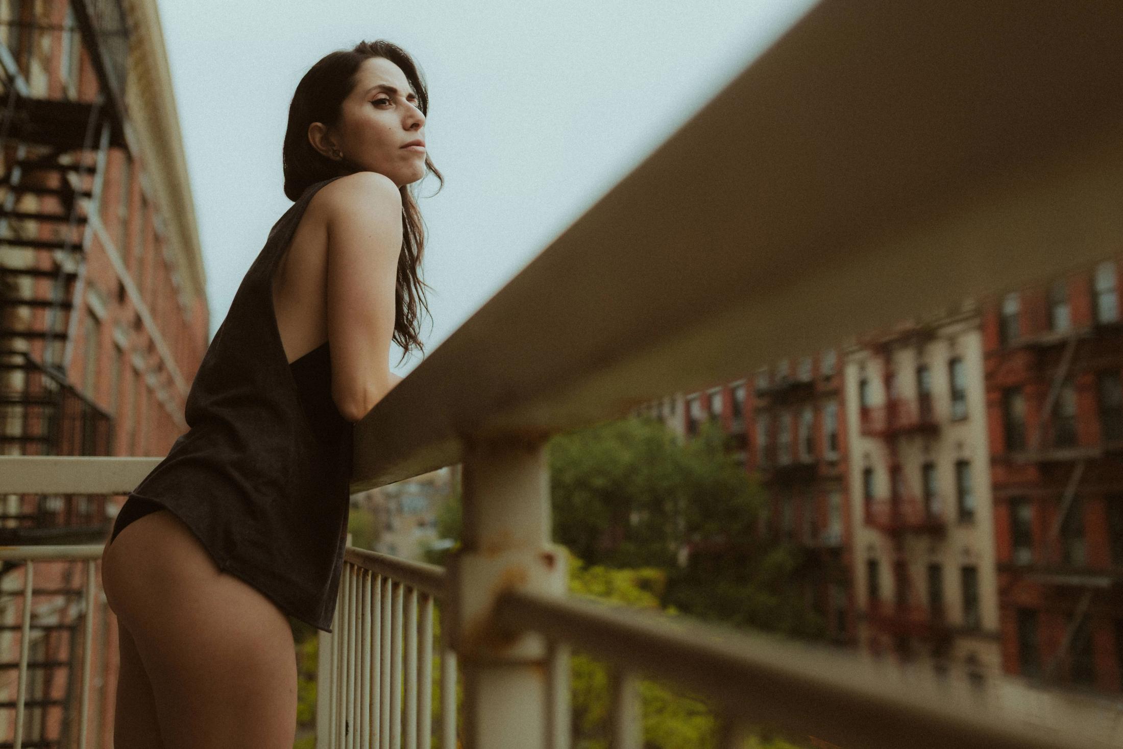Maya standing in black at the balcony railing with the city behind her.