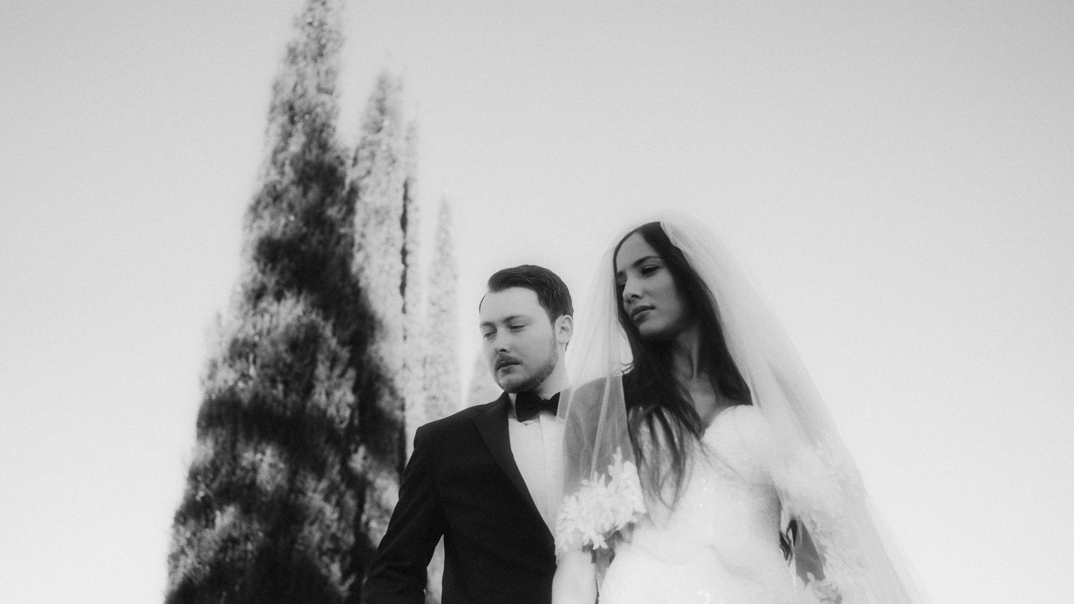 Low-angle black-and-white portrait of the couple with slender trees behind them.
