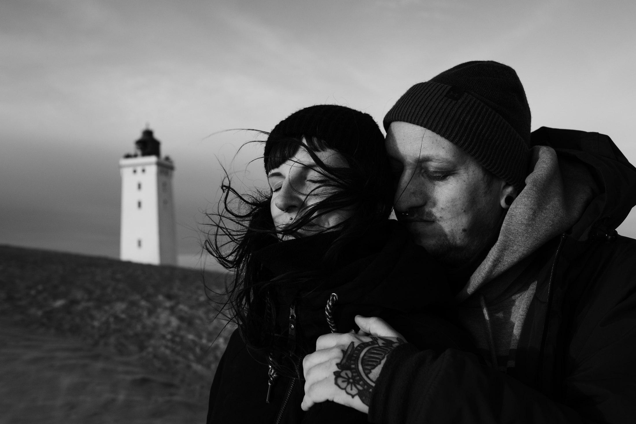 Black-and-white photograph of Jana and Bjørn embracing in the wind, with a lighthouse behind them in Denmark.