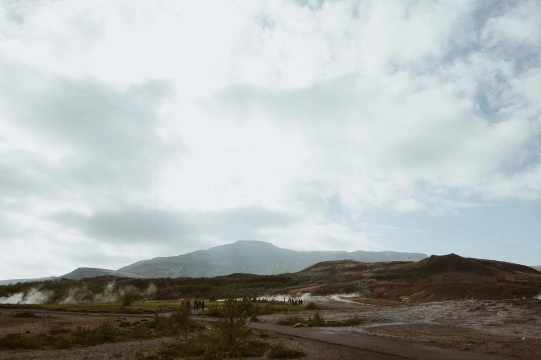 Steam rising from open geothermal ground beneath a low mountain and cloudy sky.