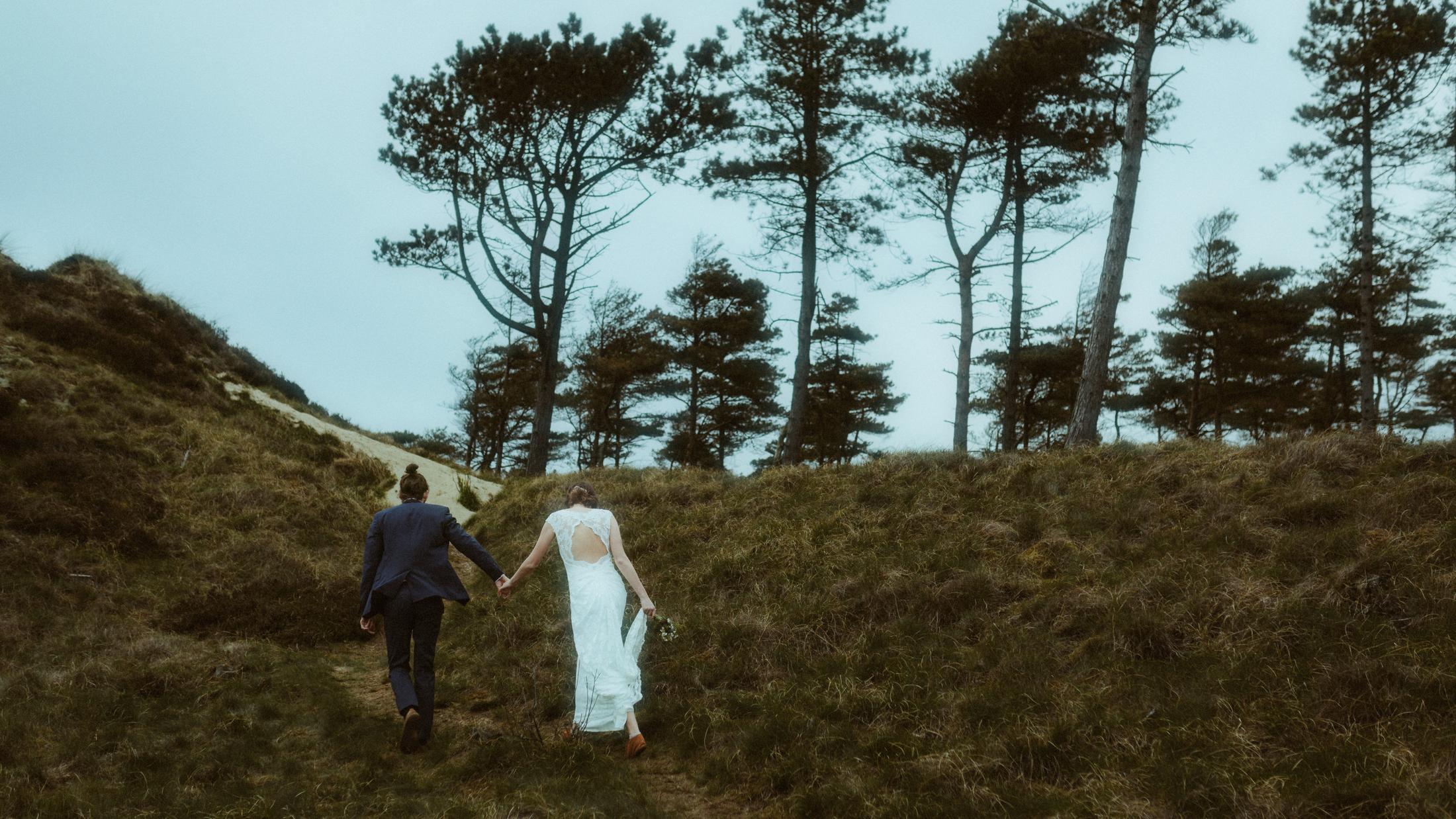 Nina und Torben laufen Hand in Hand einen grasigen Hang hinauf auf eine Reihe Kiefern zu.