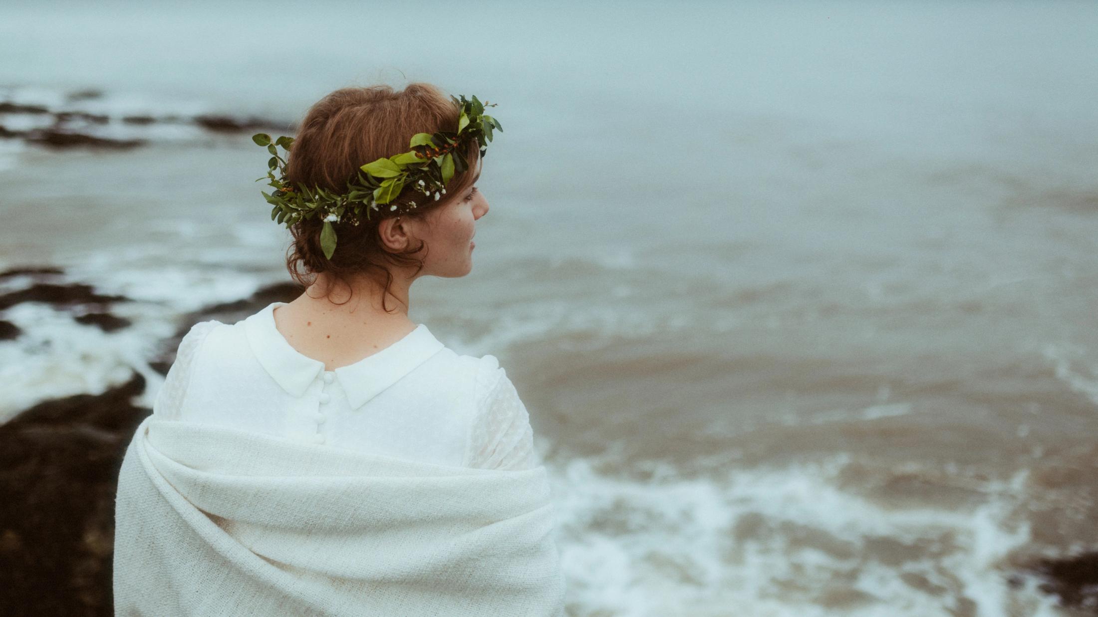 Jule von hinten in weißem Kleid und mit Blumenkranz, mit Blick über das Meer.