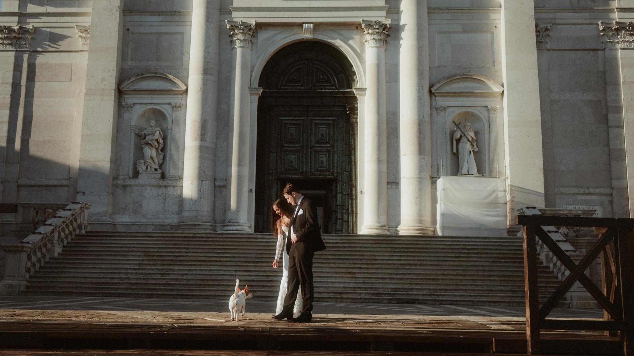The couple stands small at the foot of a monumental Venetian church, with a dog nearby.