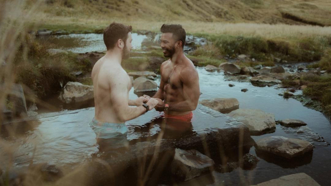 Kris and Ilya stand chest-deep in a small natural hot spring, talking face to face.