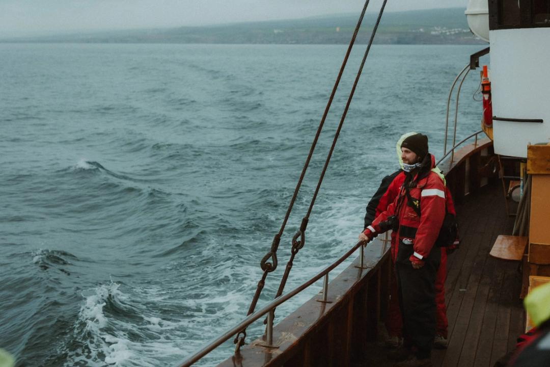 A person in a red jacket standing beside the railing of a wooden boat at sea.