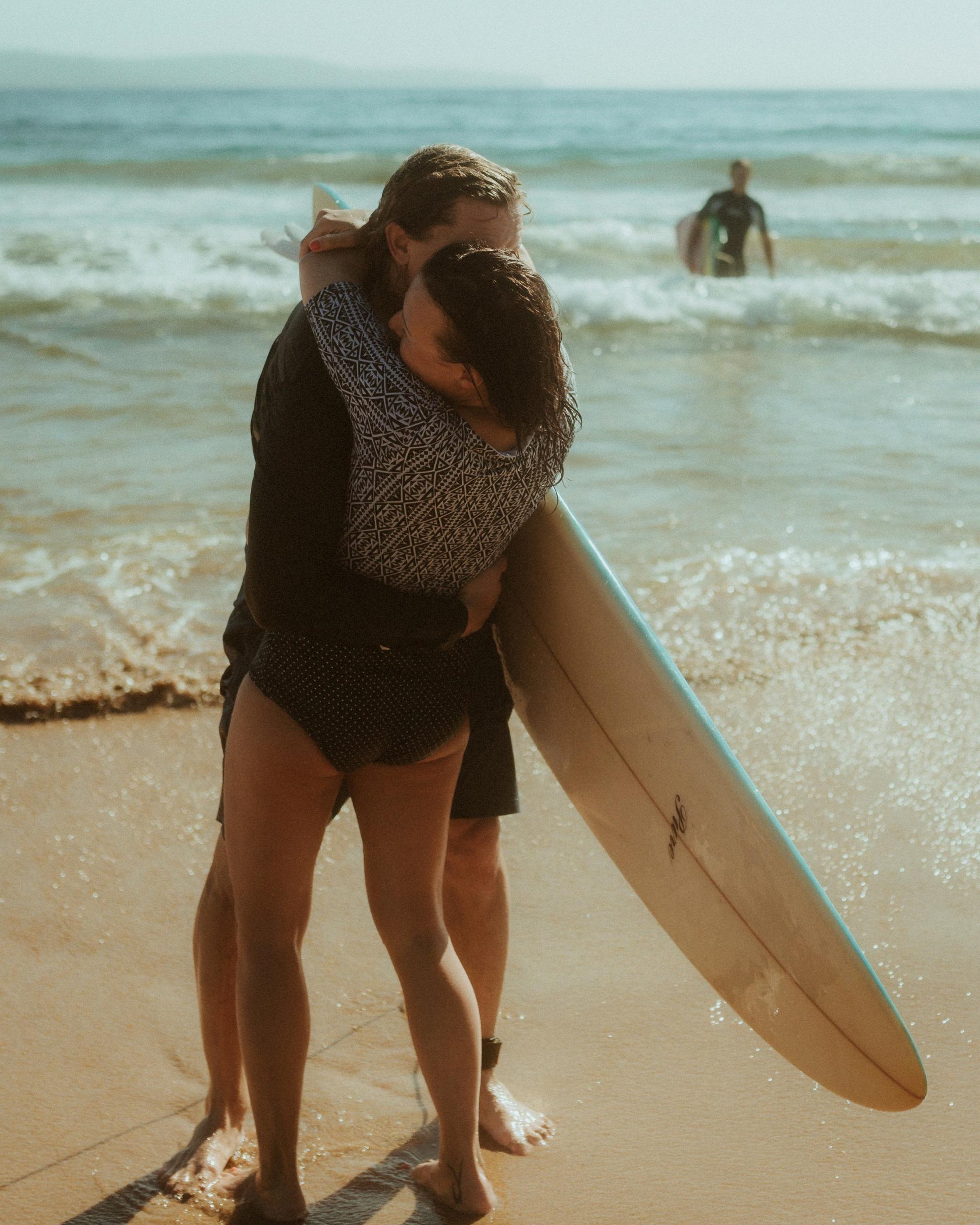 Petra and Roman embracing beside a surfboard at the water's edge