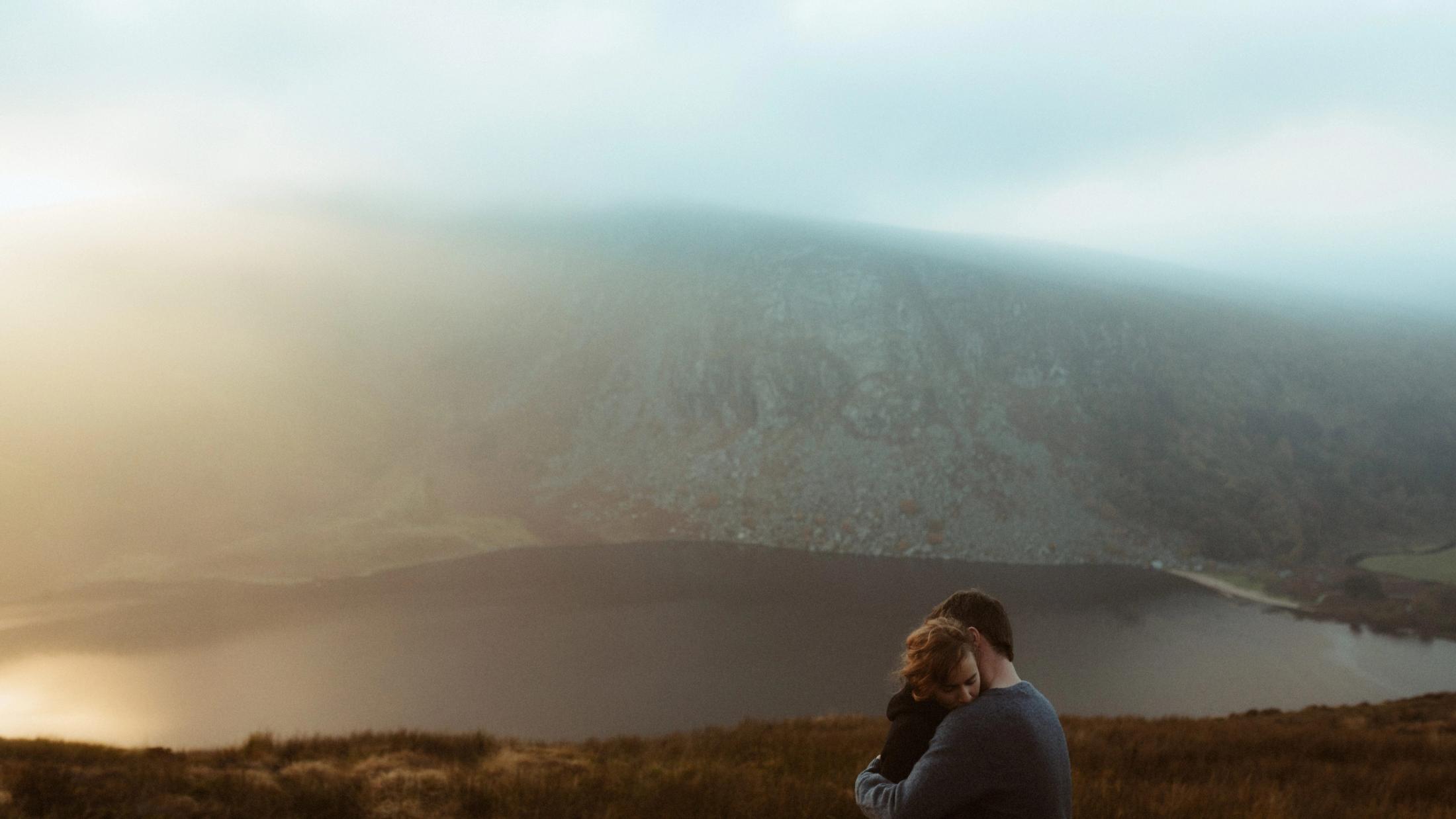 Jule und Rory umarmen sich auf einem nebligen Berggipfel über einem See und Tal.