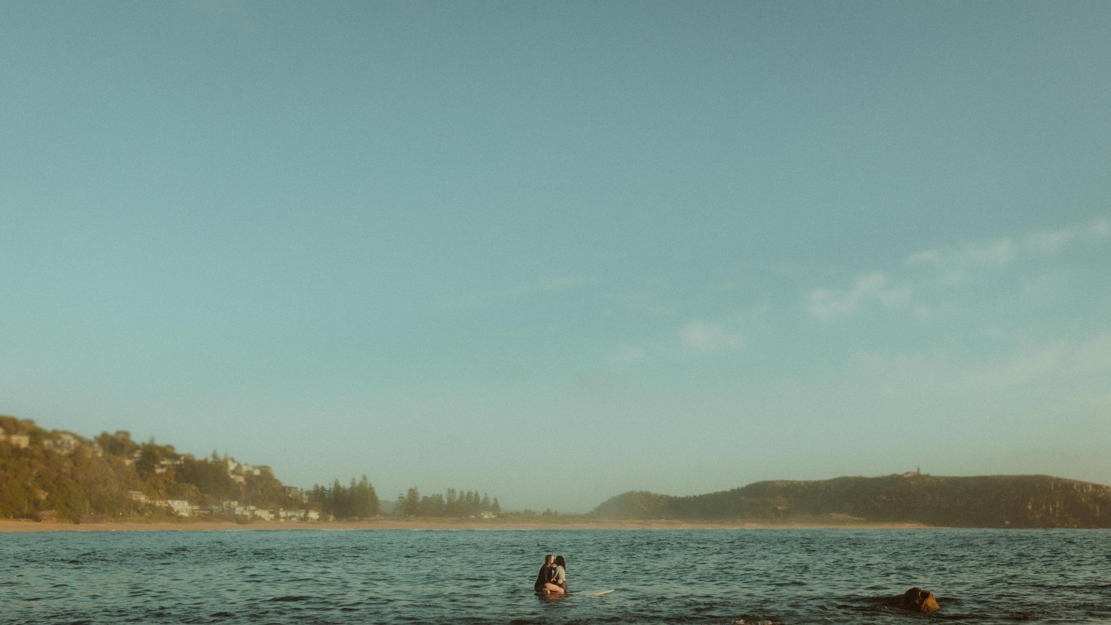 Petra and Roman appearing small on a surfboard beneath a wide morning sky