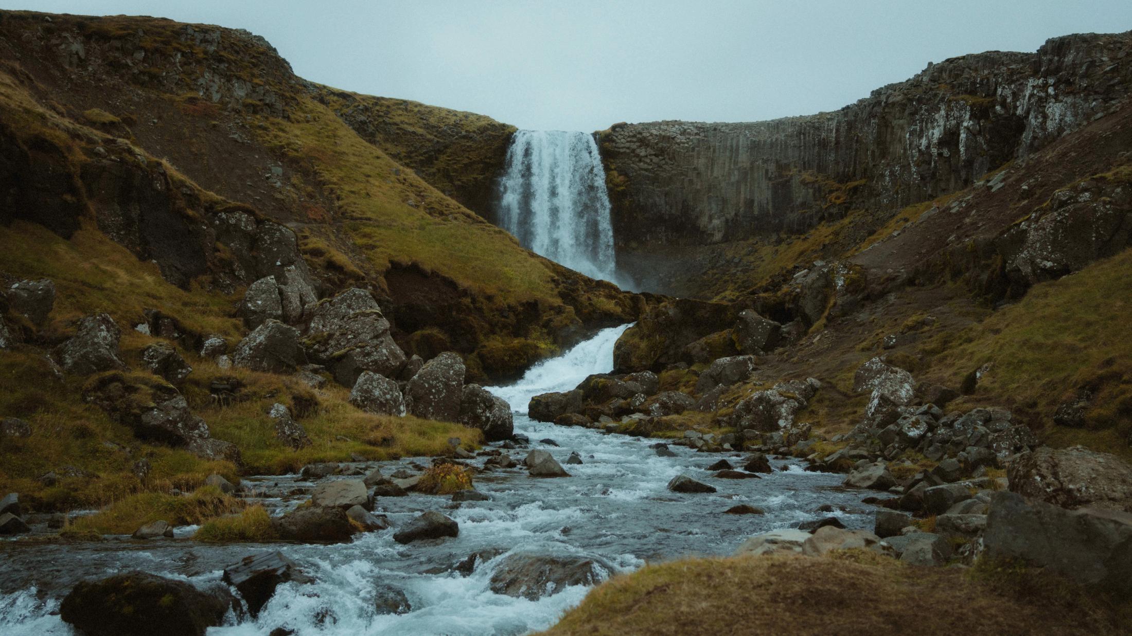 Ein breiter Wasserfall fällt zwischen moosigen Hängen in einen felsigen Bach.