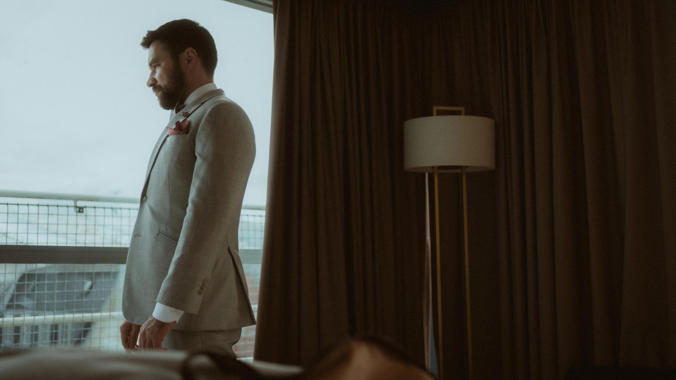 Ilya stands fully dressed in a light suit beside the hotel window.