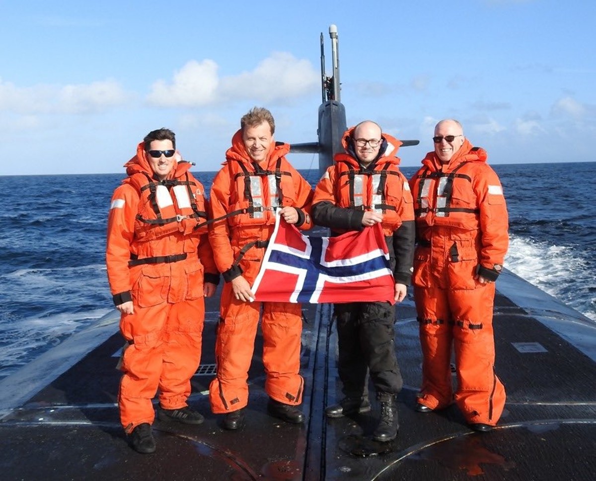 Fire menn i oransje flytedrakter holder det norske flagget mellom seg på dekket til en ubåt midt i havet