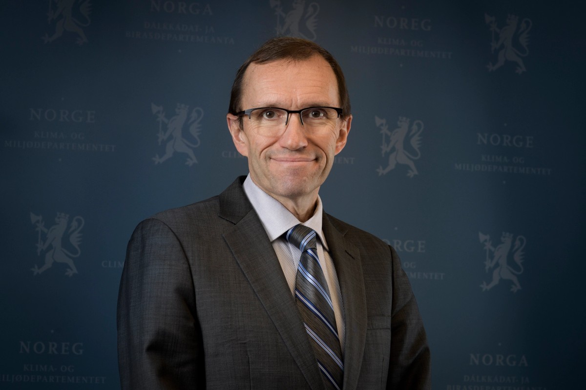 Espen Barth Eide smiler, kledd i en mørk dress for en nøytral bakgrunn med regjeringens logo