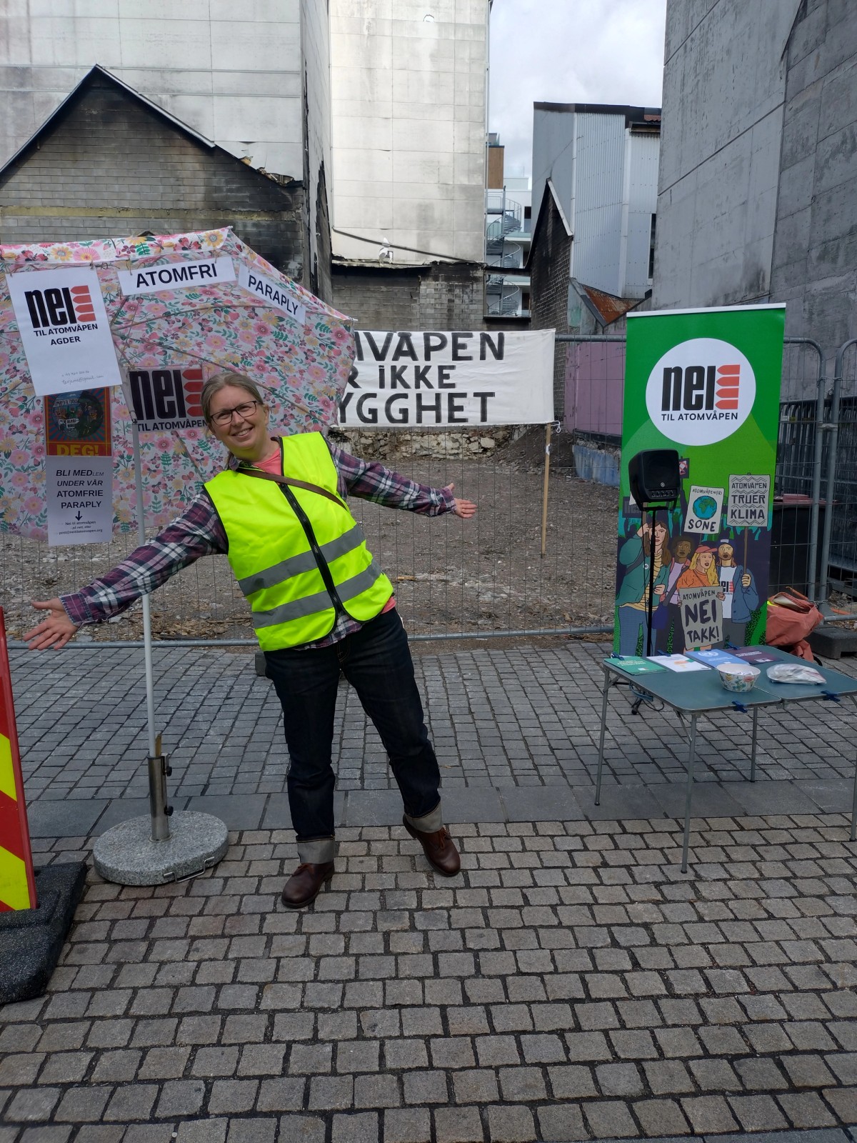En aktivist står på stand. Hun har på seg gul refleksvest og smiler foran en blomstrete paraply