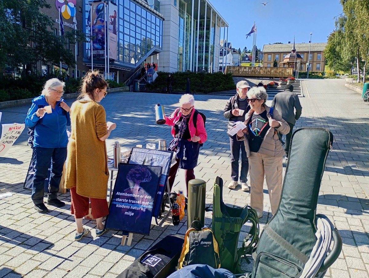Folk står rundt et bord med kaffekanner og kopper i Tromsøs gågate. Det er strålende solskinn, og folkene smiler. Ved siden av bordet er plakater om atomvåpentrusselen.