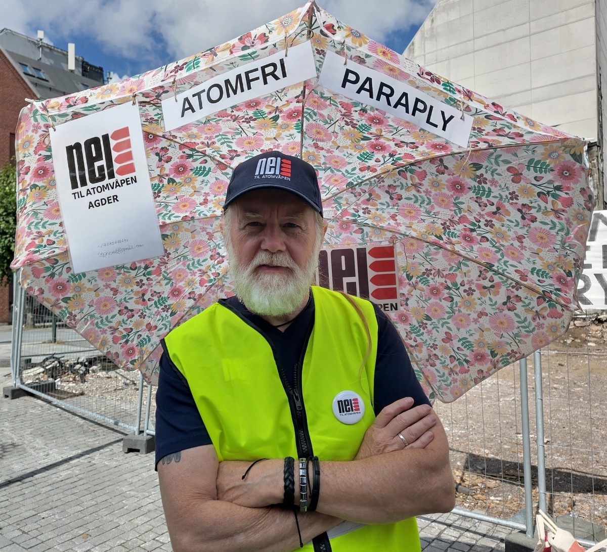 En mann står foran en blomstrede paraply med lappene "atomfri paraply" og "nei til atomvåpen agder". Han har refleksvest og caps på, der det står "Nei til Atomvåpen.