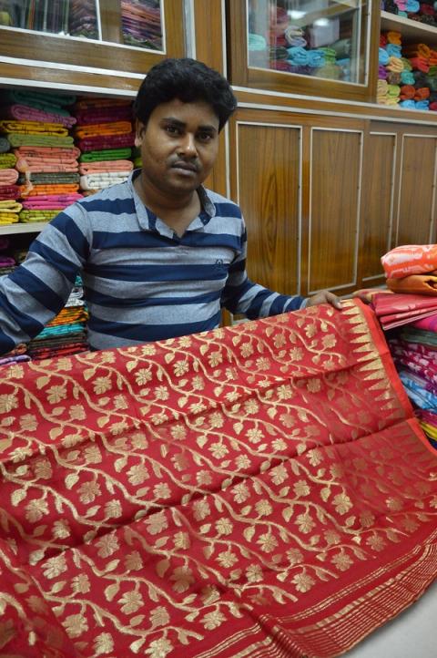 A shopkeeper displays a red tussar silk saree with gold floral motifs