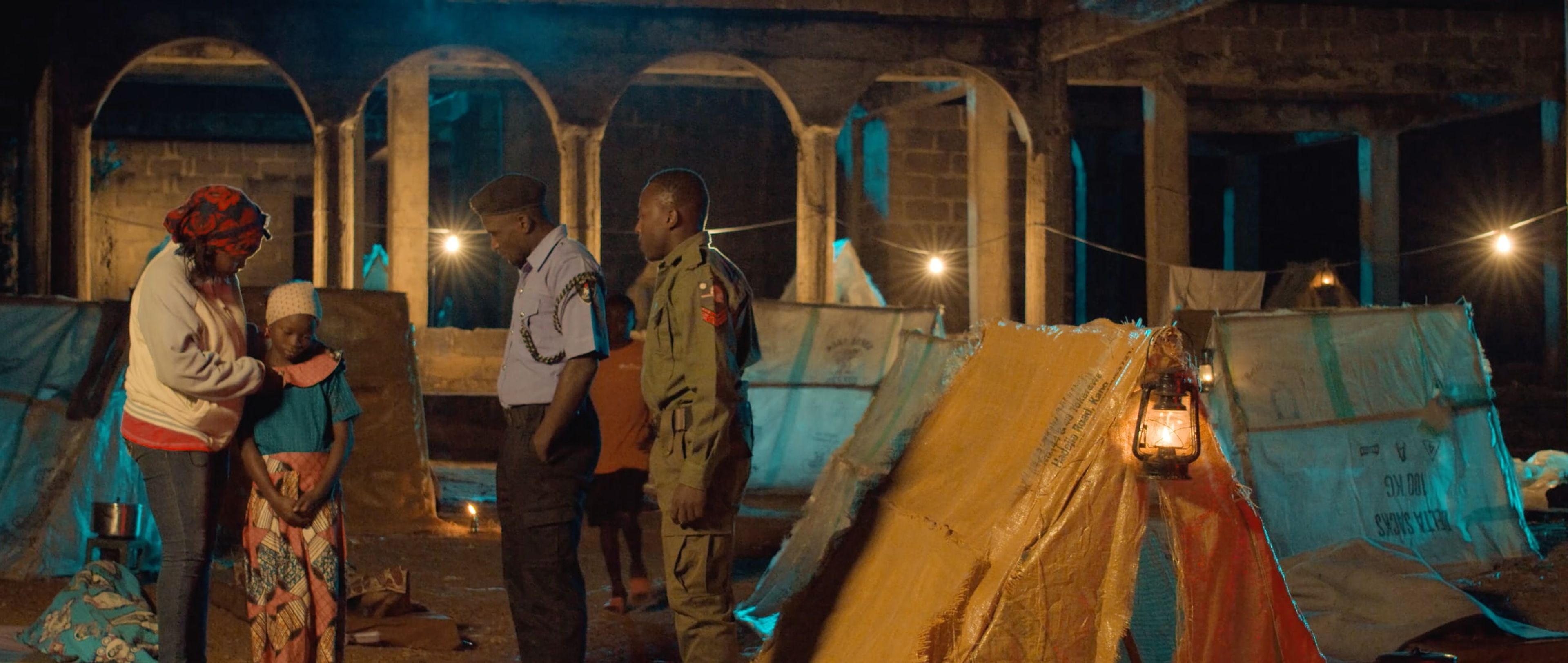 Travellers and soldiers among lantern-lit tents inside a ruined stone building at night