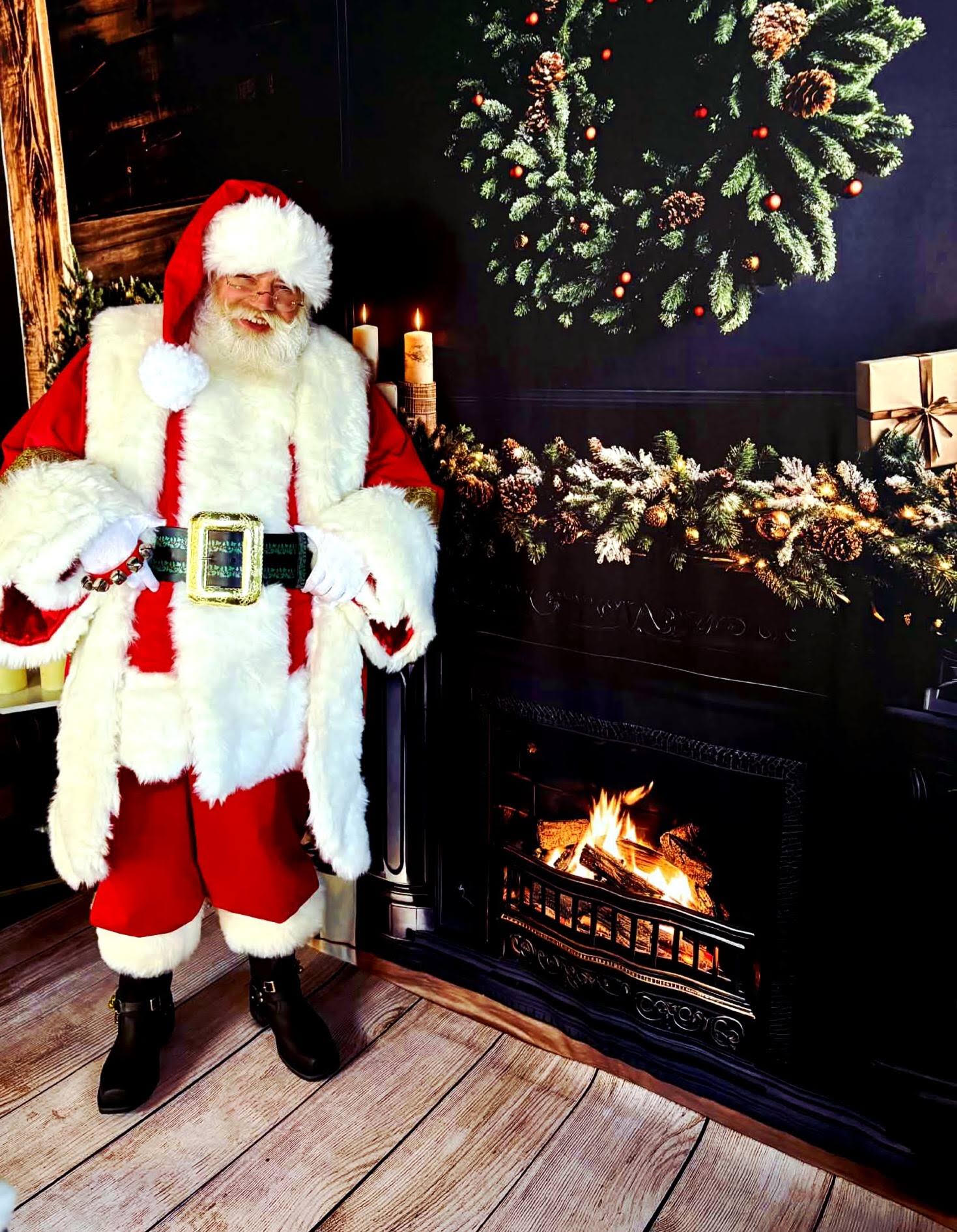Santa Claus in a red and white suit beside a fireplace and wreath