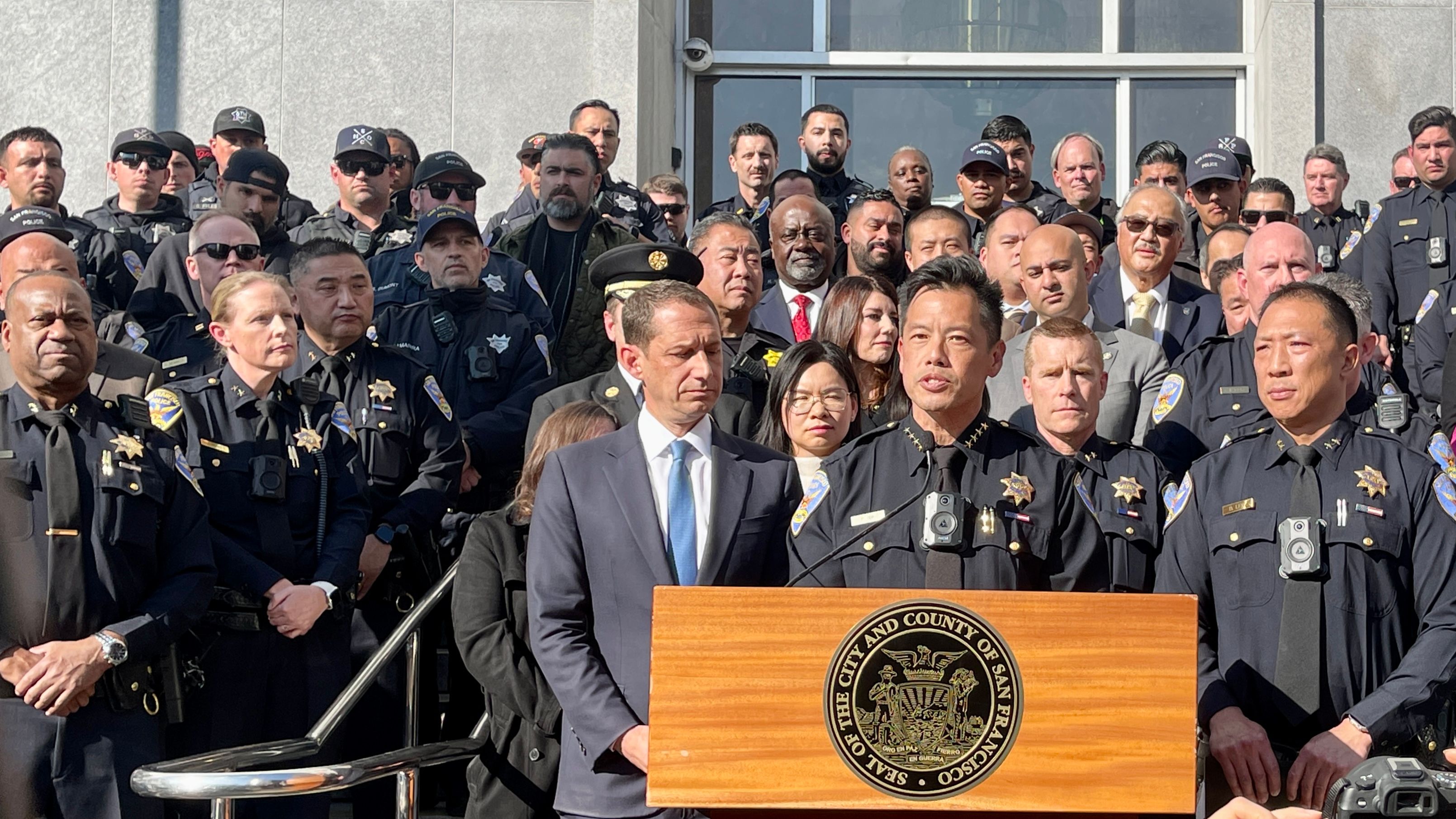 Mayor Daniel Lurie (left in front row) thanks the service and leadership of Interim Police Chief Paul Yep (speaking) and introduces his appointment of incoming Police Chief Derrick Lew (right in front row) on the steps outside Hall of Justice where also houses the San Francisco Police Department’s Investigation Bureau. Photo by Portia Li