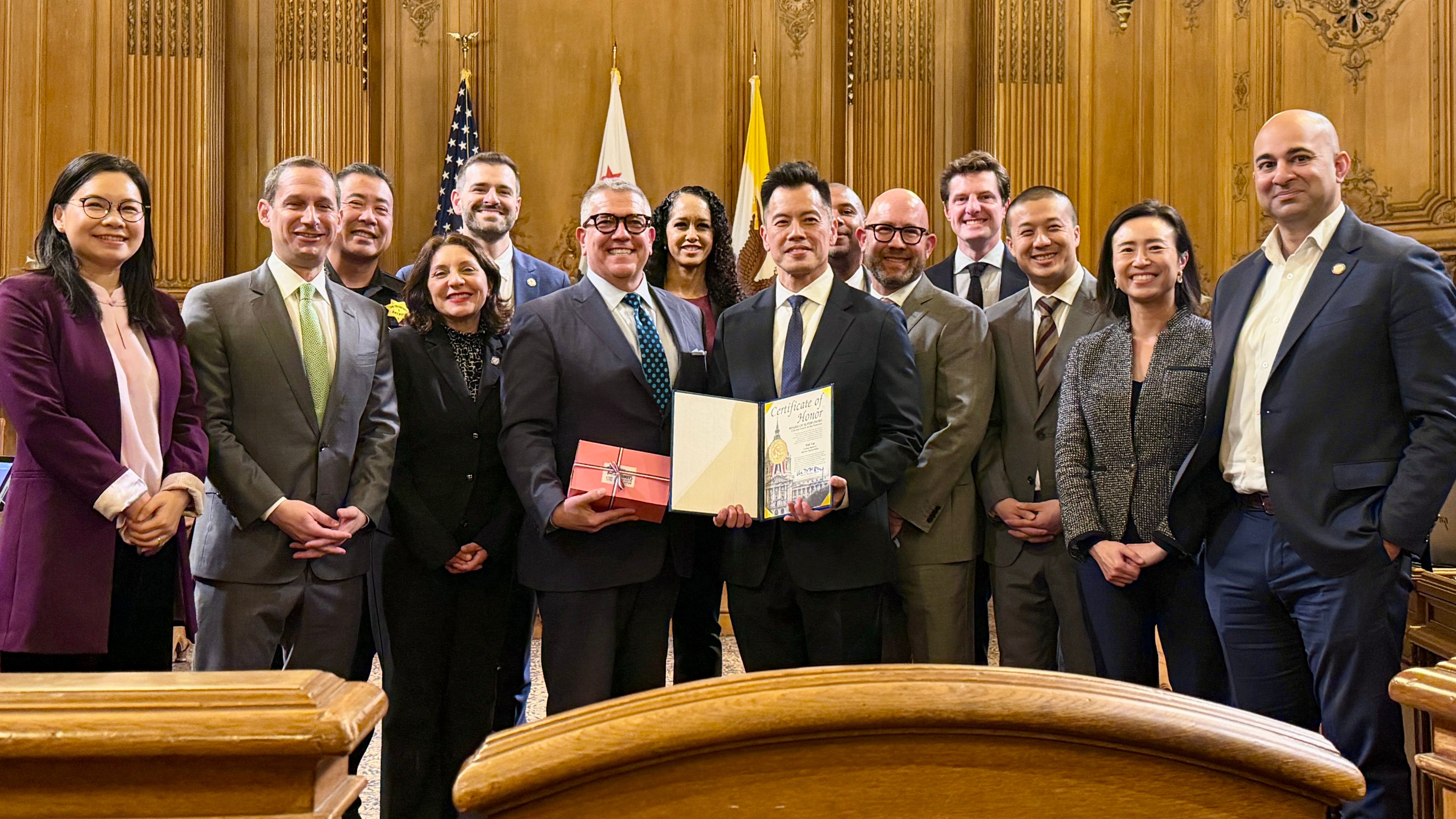 Outgoing Interim Police Chief Paul Yep (center right in front row) is recognized by the Board of Supervisors for his leadership and accomplishment to lead the San Francisco Police Department from June to December 2025 with record low crime rates and high police hiring. District 6 Supervisor Matt Dorsey (center left in front row) introduces the Recognition of Commendation to honor Yep. Mayor Daniel Lurie joins the ceremony (2nd from far left). Photo by Portia Li