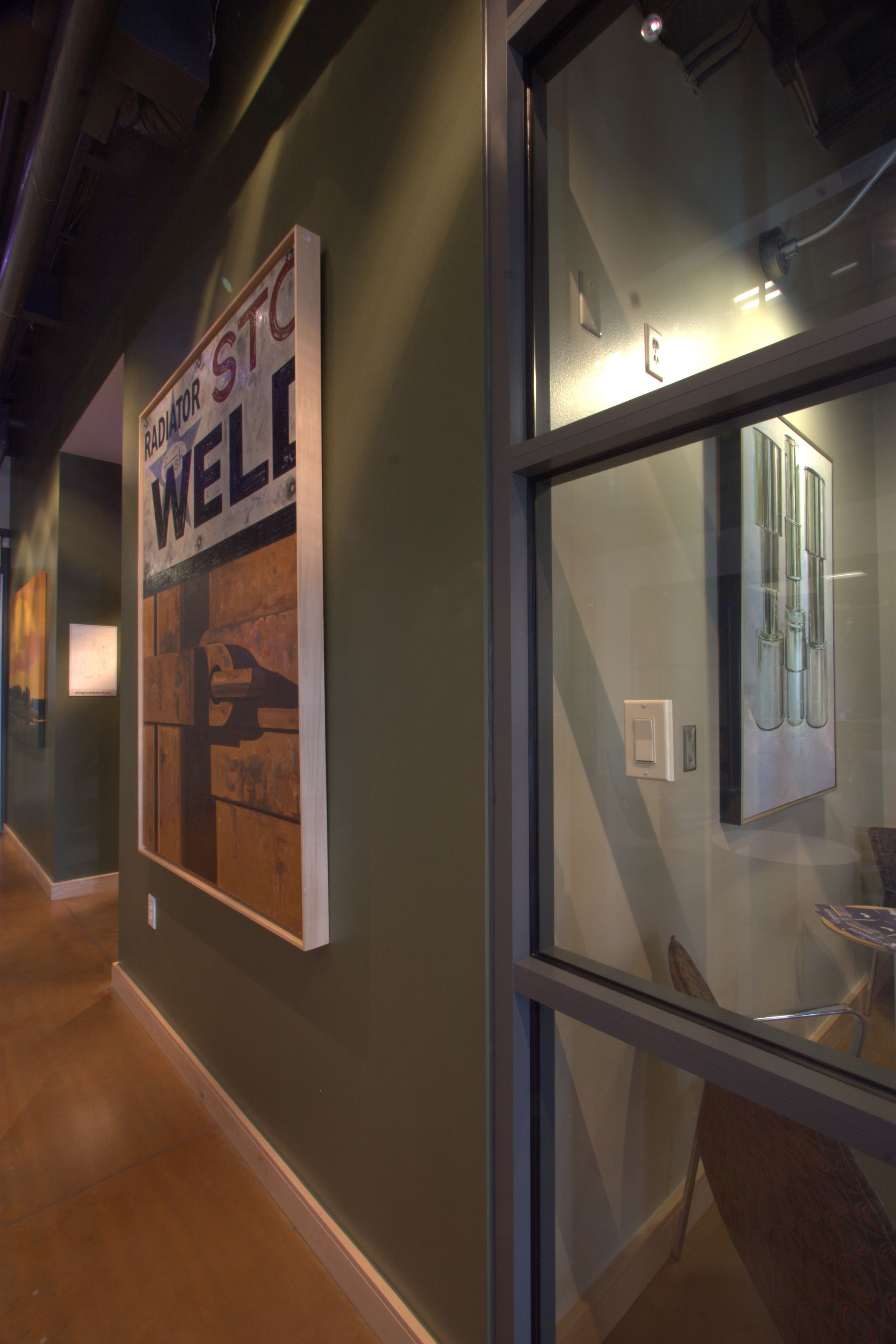 A hallway with a dark green wall displaying a large "Radiator Stop Well" sign artwork, a polished brown floor, and a glass partition to an adjacent room.