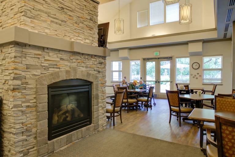 Gardens at Columbine Memory Care living room with a stone fireplace, dining tables, and patio doors