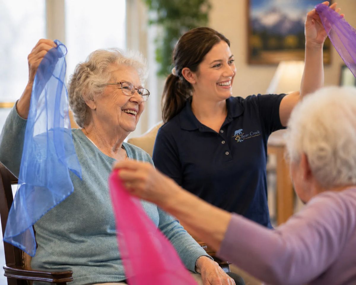 A laughing Bear Creek resident raises a blue chiffon scarf overhead during a seated movement class led by a staff member in a Bear Creek polo, with another resident waving a pink scarf nearby