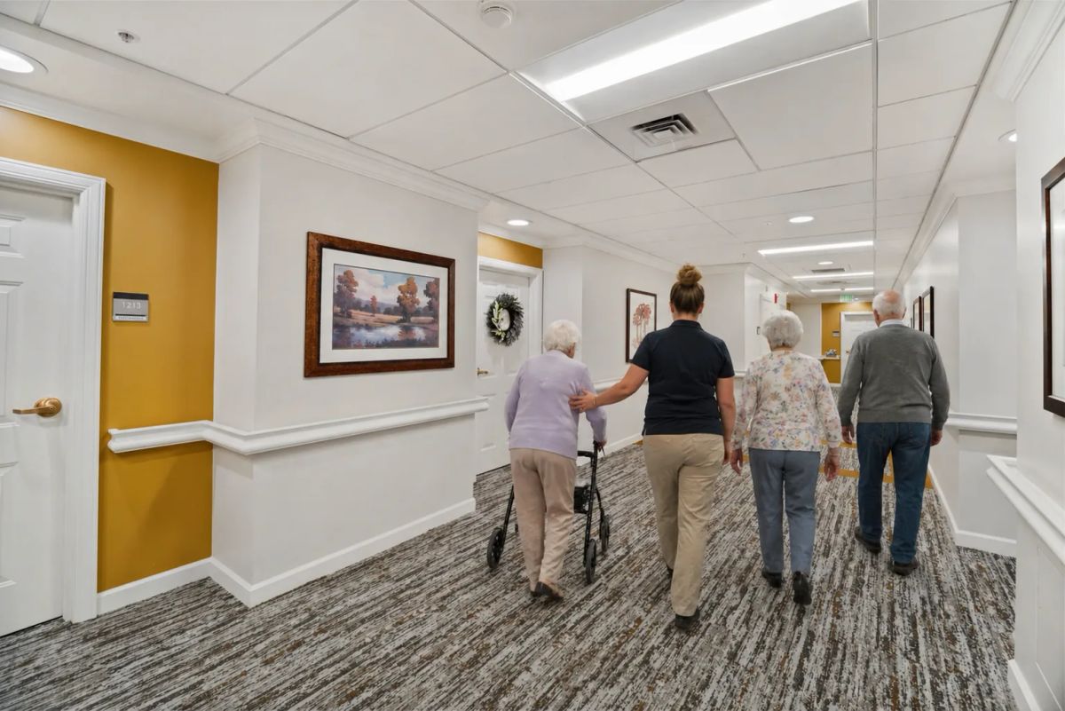 A Bear Creek staff member walks down a remodeled corridor alongside three residents, including one using a rollator, past framed art and a yellow accent wall