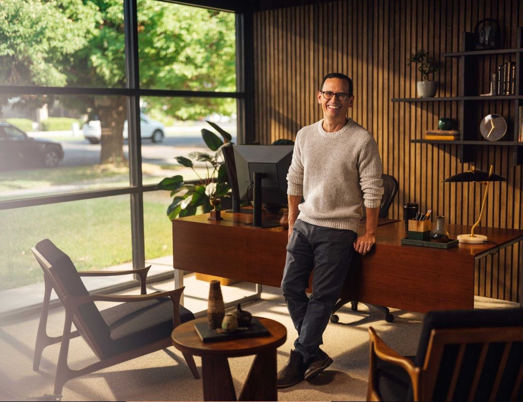 A photo of a man leaning against his desk, smiling. 