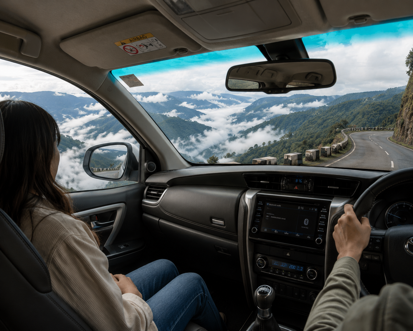 Interior view of a touring vehicle driving along a high mountain cliff road in Meghalaya with a passenger looking out at a deep green river valley filled with rolling white clouds