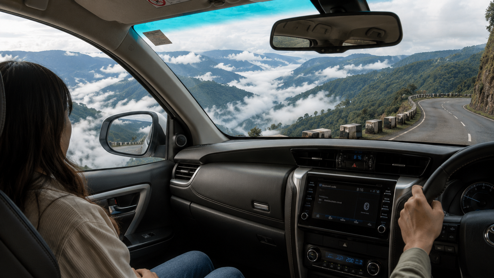 Interior view of a touring vehicle driving along a high mountain cliff road in Meghalaya with a passenger looking out at a deep green river valley filled with rolling white clouds