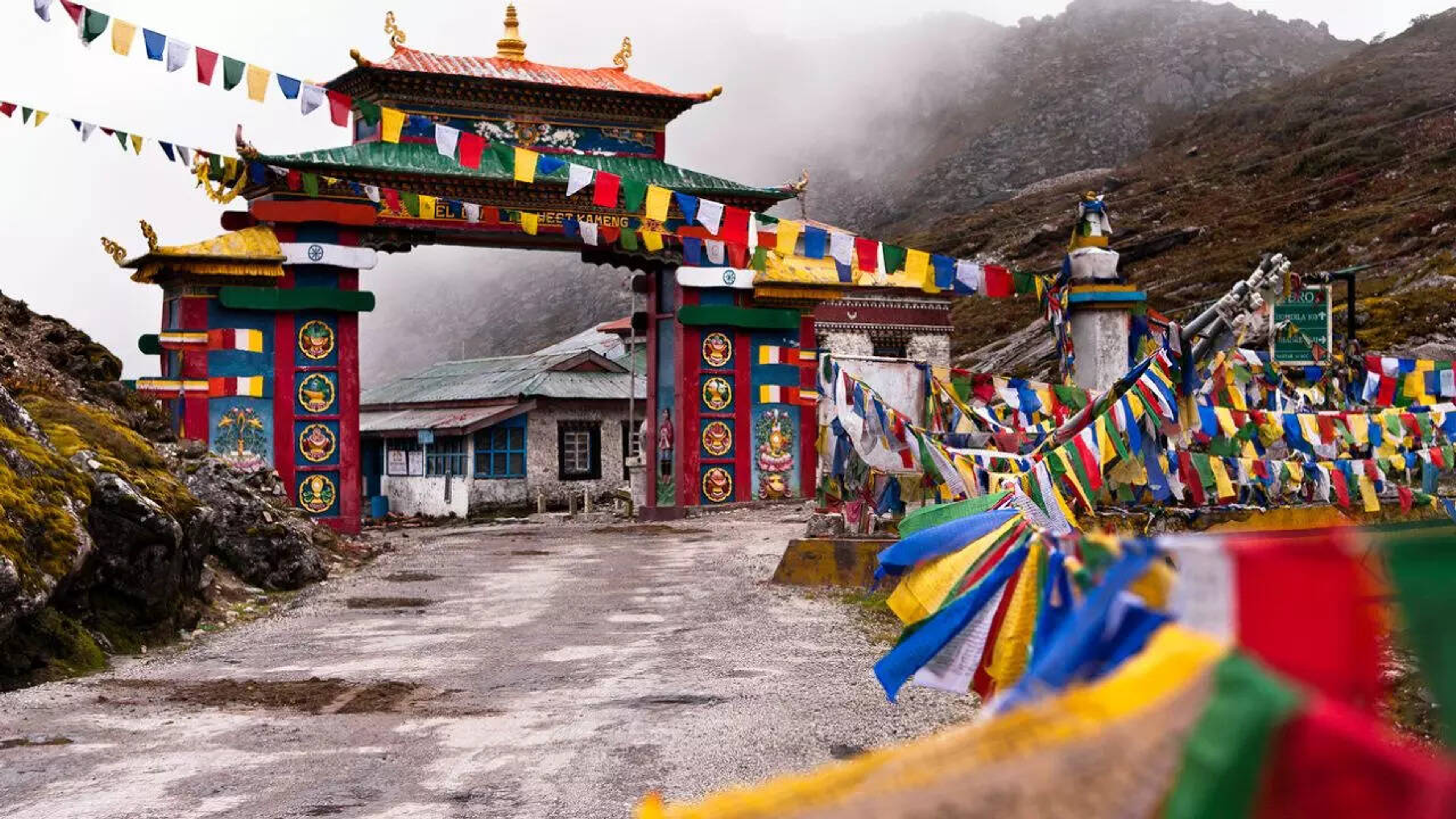 The colorful Tawang Welcome Gate adorned with vibrant red, blue, and white prayer flags against a backdrop of snow-capped mountains in Arunachal Pradesh.