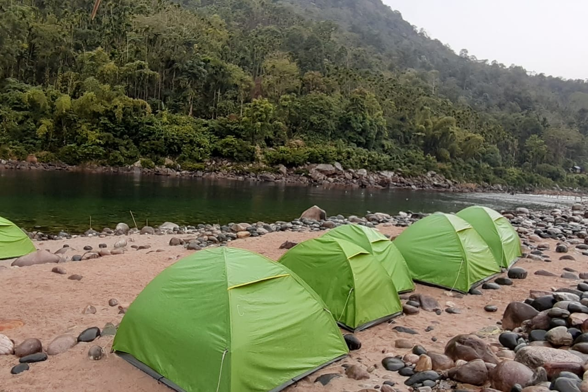 Premium white adventure tents pitched on a clean stone riverbank beside the clear Umngot River in Shnongpdeng at twilight."